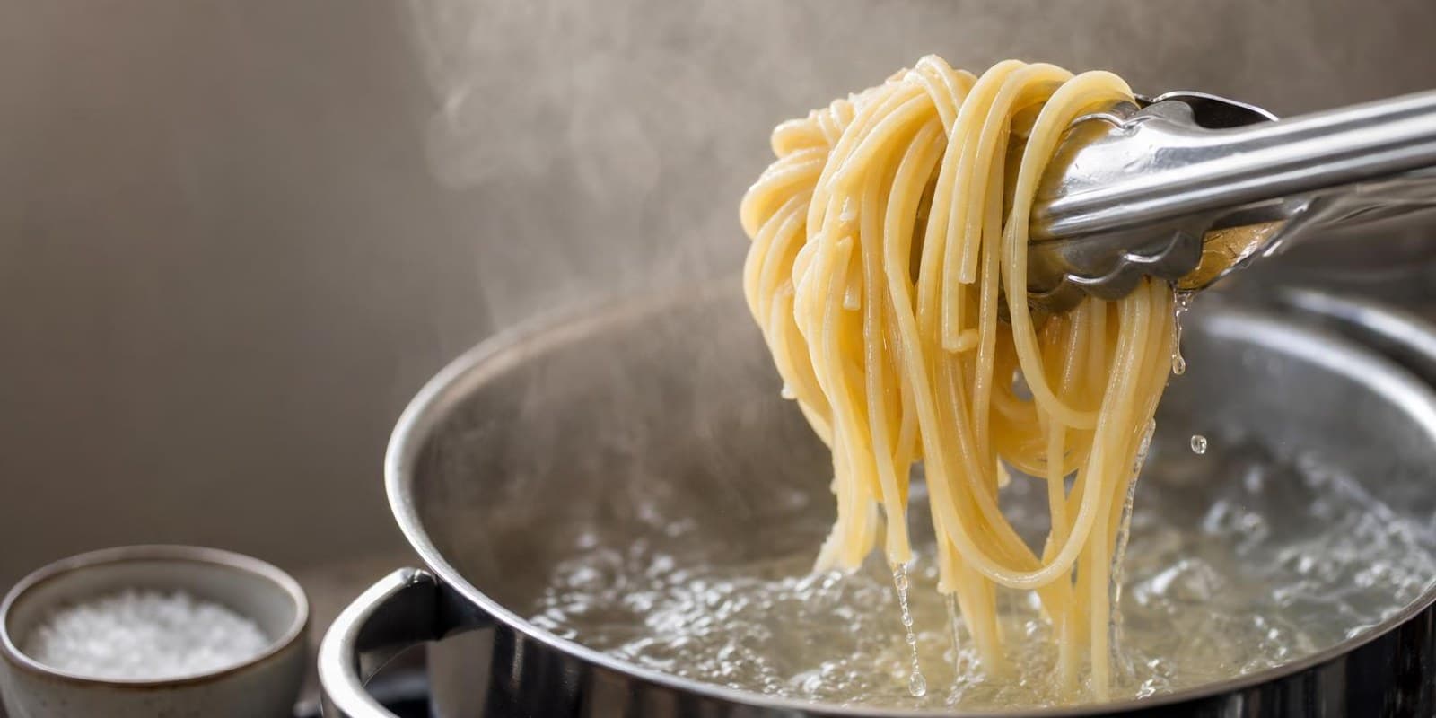 Eine Zange hebt glaenzende Spaghetti aus sprudelnd kochendem Salzwasser, Dampf steigt auf.