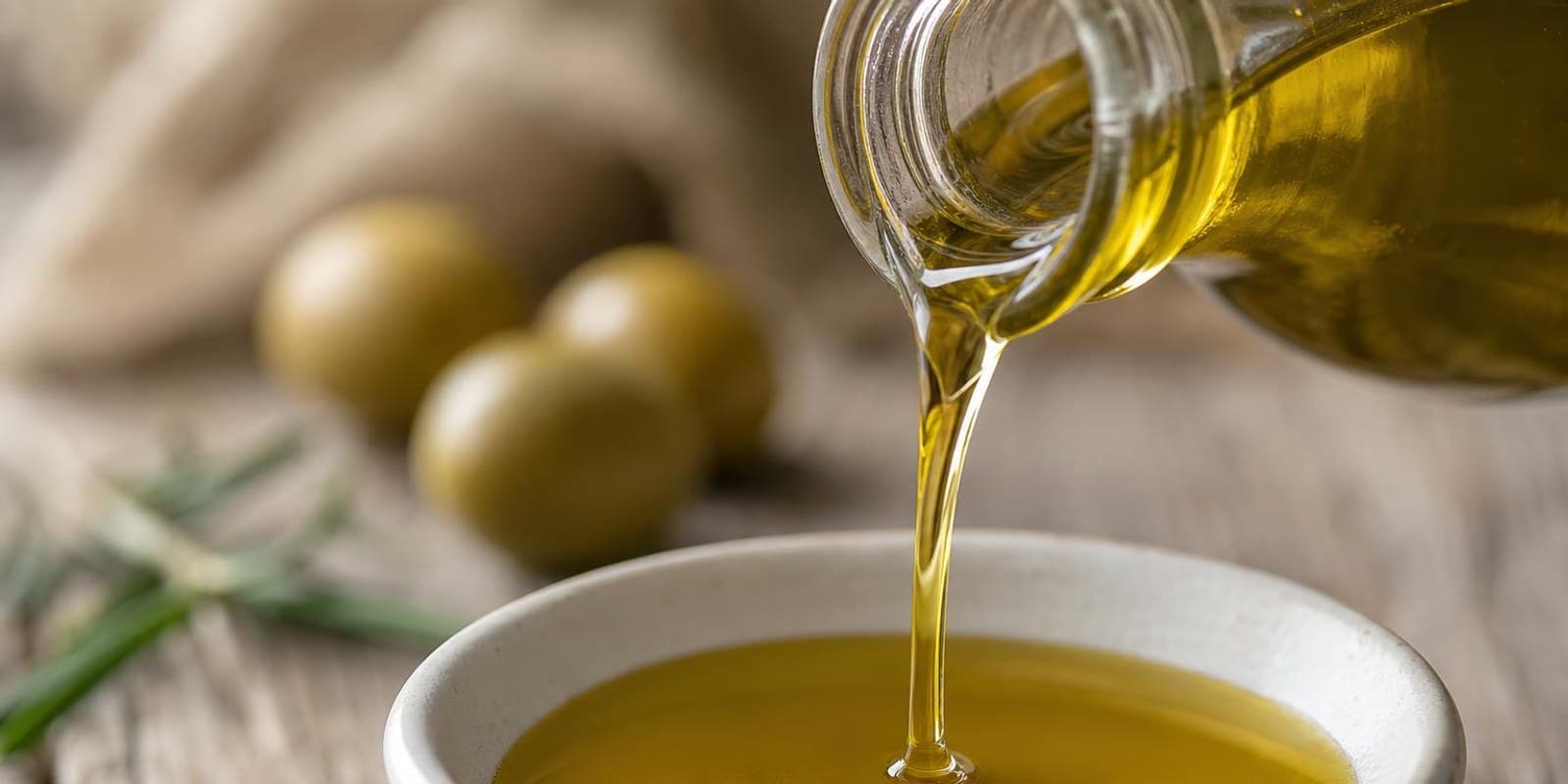 Olivenoel fliesst in eine kleine weisse Schale, daneben Oliven und Rosmarin.