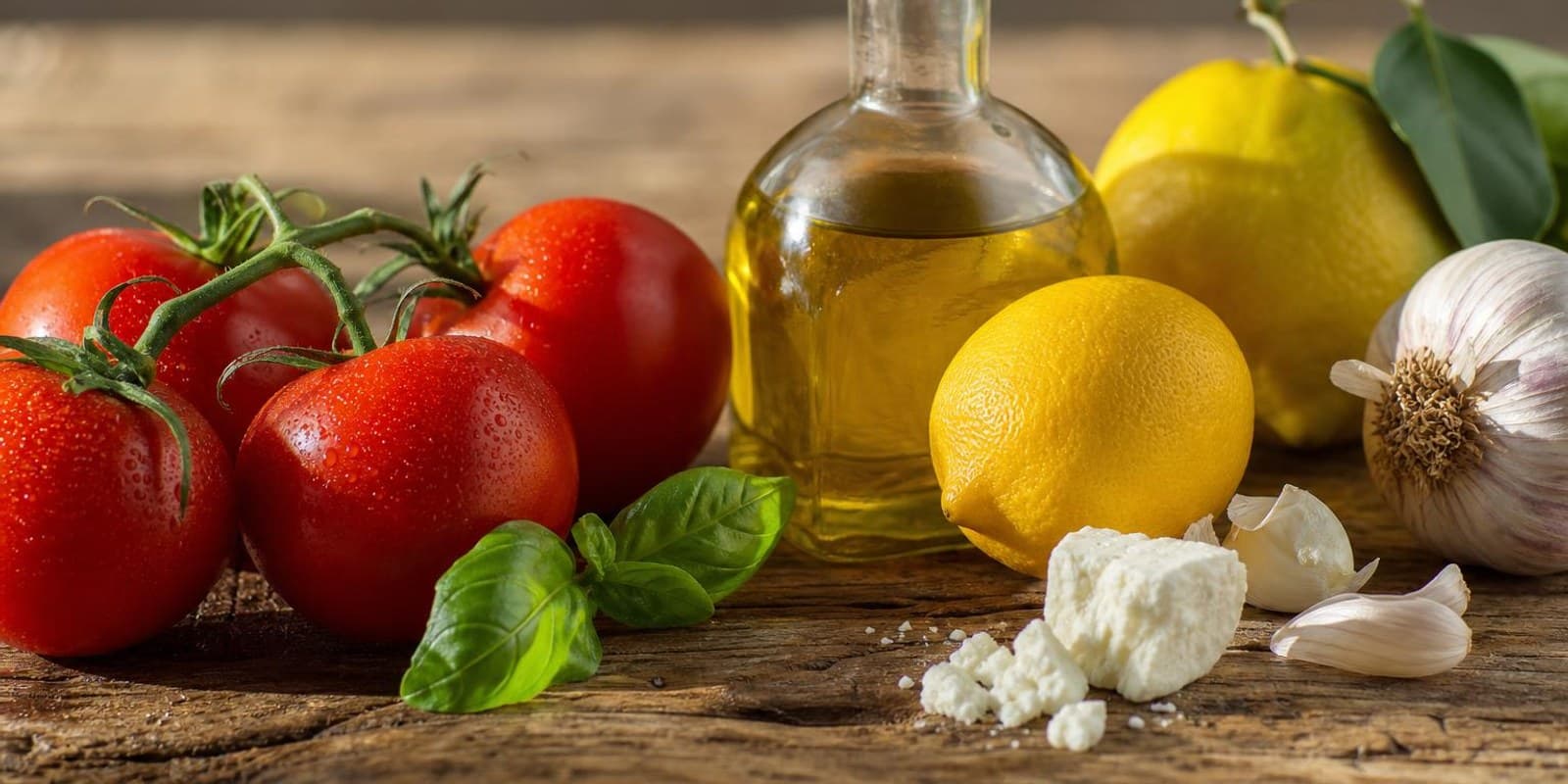 Tomaten, Olivenoel, Zitronen und Kraeuter liegen auf einem sonnenbeschienenen Holztisch.