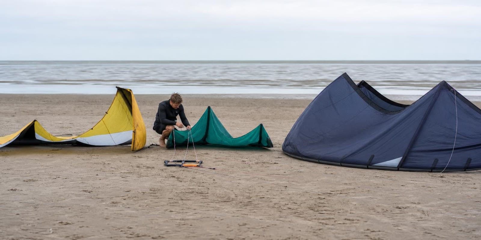 Drei unterschiedlich grosse Kites liegen nebeneinander am Strand, ein Kiter prueft die Leinen.