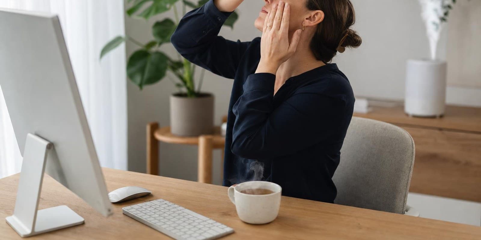 Eine Frau traeufelt sich am Schreibtisch entspannt einen Augentropfen ins Auge, daneben dampft eine Teetasse.