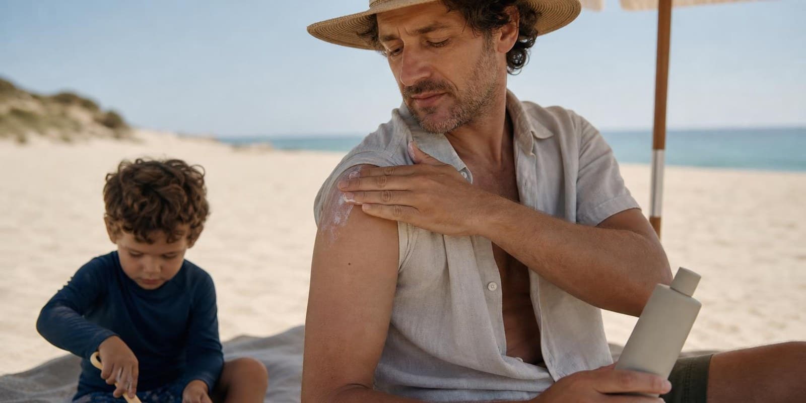 Mann cremt sich im Schatten eines Sonnenschirms am Strand mit Sonnencreme ein.