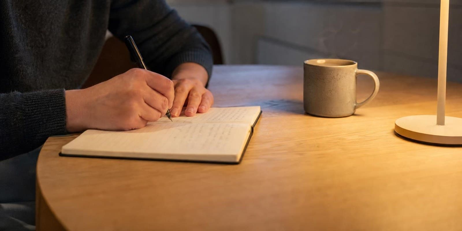 Haende schreiben abends bei Lampenlicht in ein einfaches Notizbuch, daneben steht eine Tasse Tee.