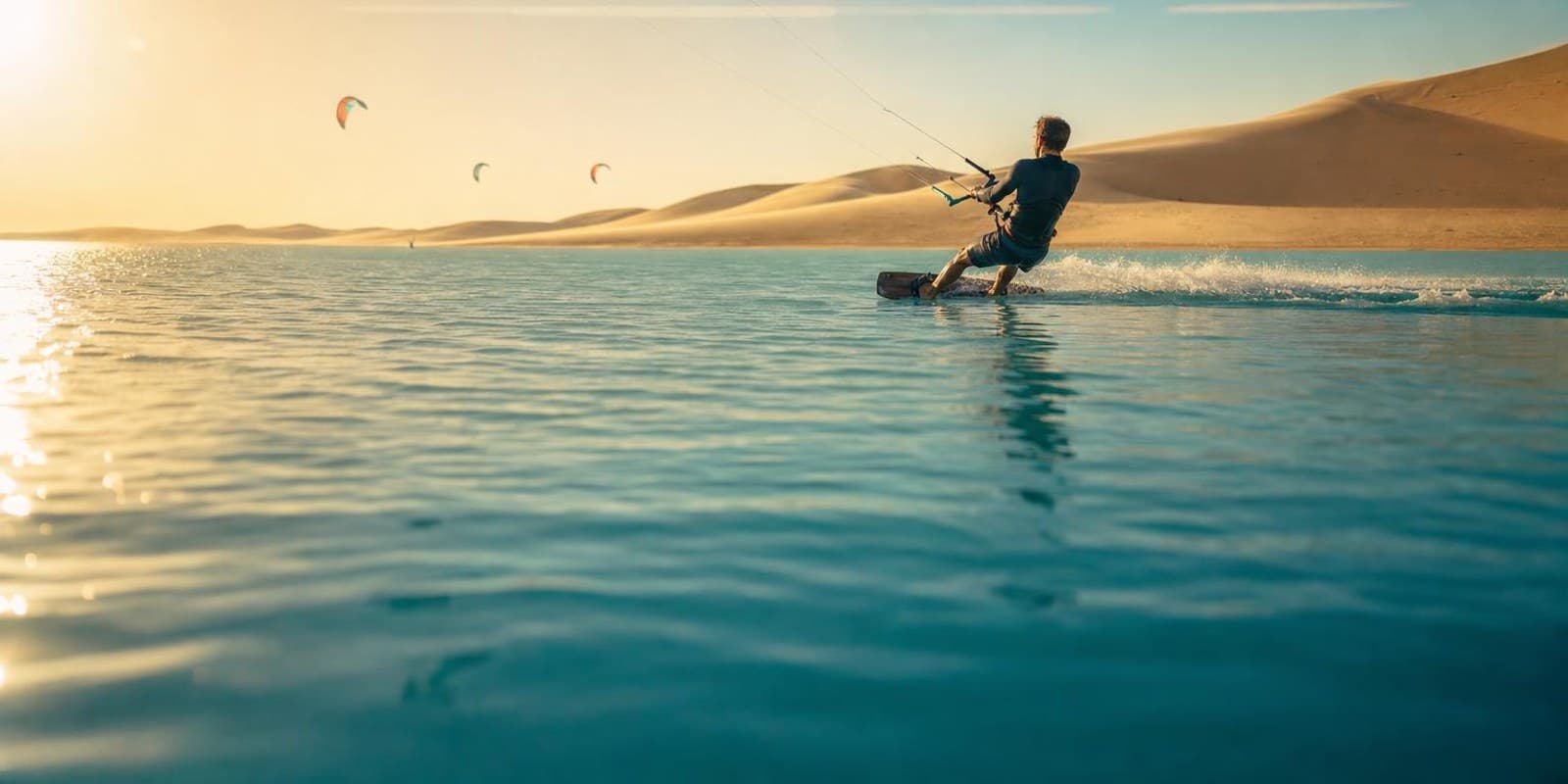 Kitesurfer gleiten ueber eine flache tuerkise Lagune, dahinter leuchten die Sandduenen der Sahara.