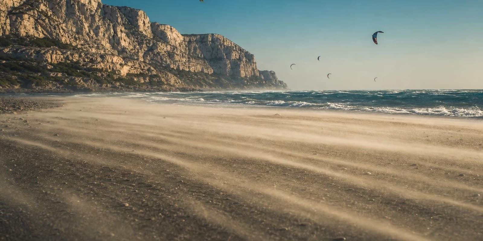 Sturmgepeitschter Strand bei Leucate, Sand weht flach ueber den Boden und viele Kites rasen ueber Schaumkronen.