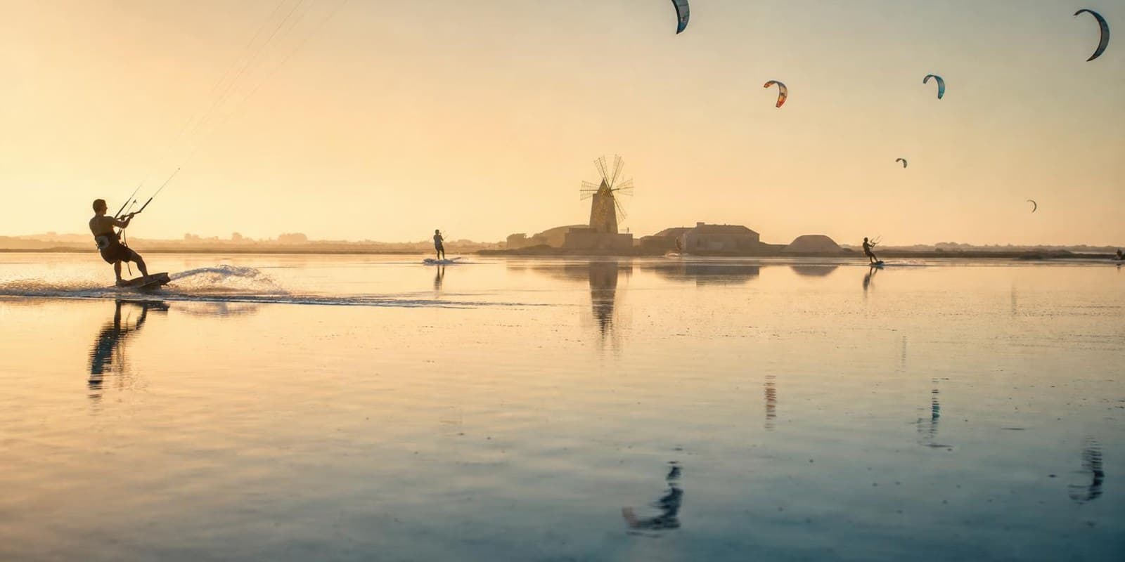 Kniehohe Lagune auf Sizilien mit Kitesurfern, am Ufer die Silhouette einer alten Windmuehle.