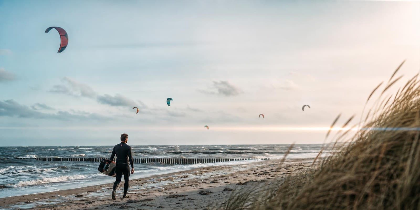 Kitesurfer traegt sein Board am Ostseestrand mit Buhnen entlang, Kites am Himmel.