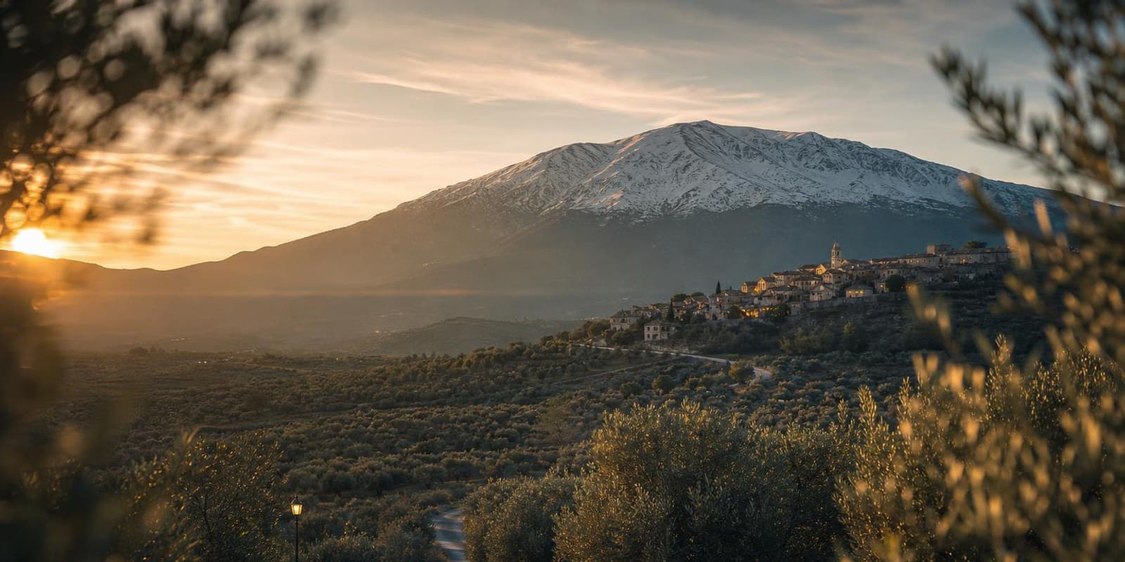 Strasse fuehrt durch Olivenhaine auf einen hohen griechischen Berg zu, ein Dorf liegt am Hang.