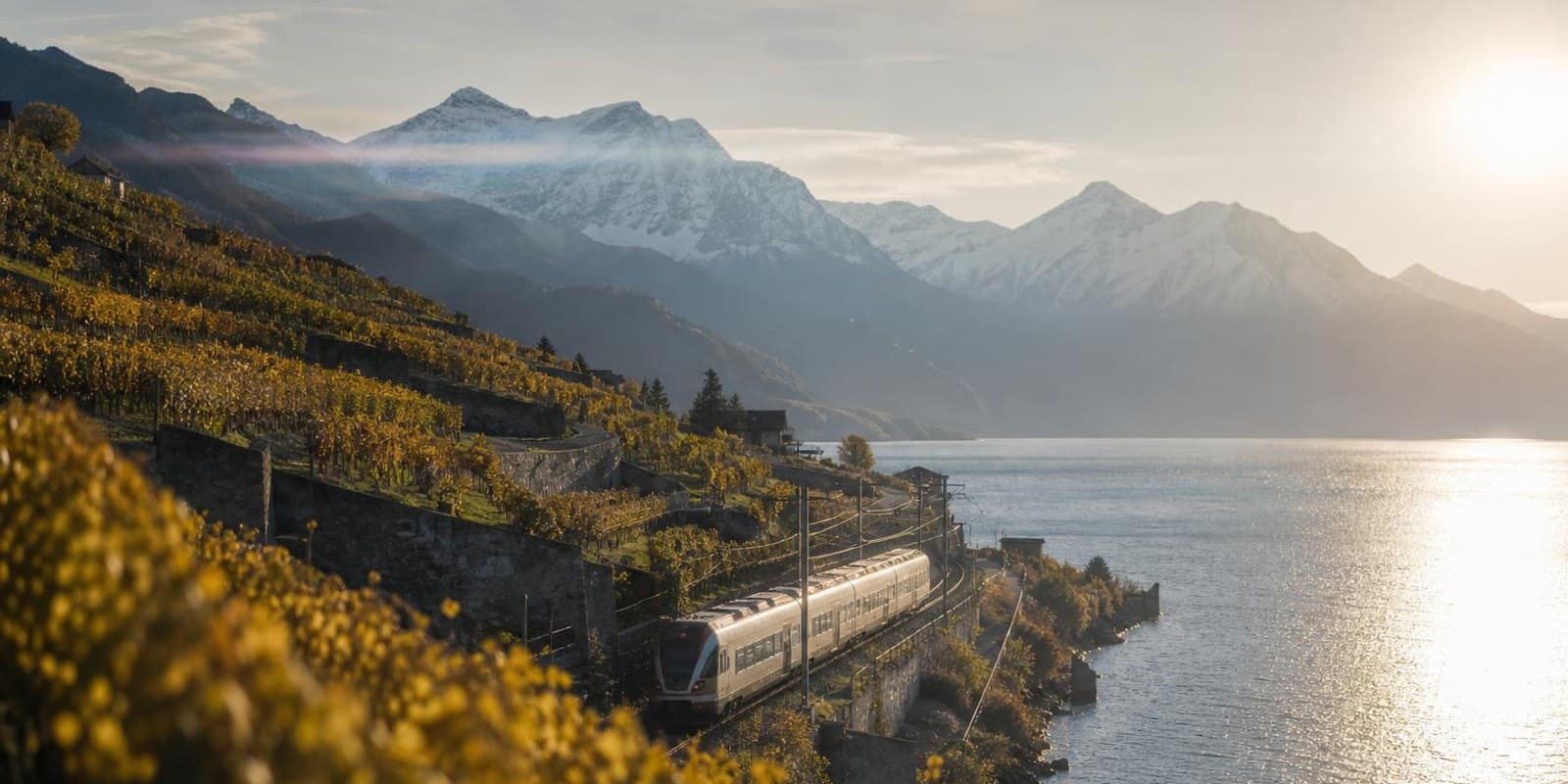 Zug faehrt am Ufer eines Schweizer Sees entlang, dahinter schneebedeckte Alpengipfel.