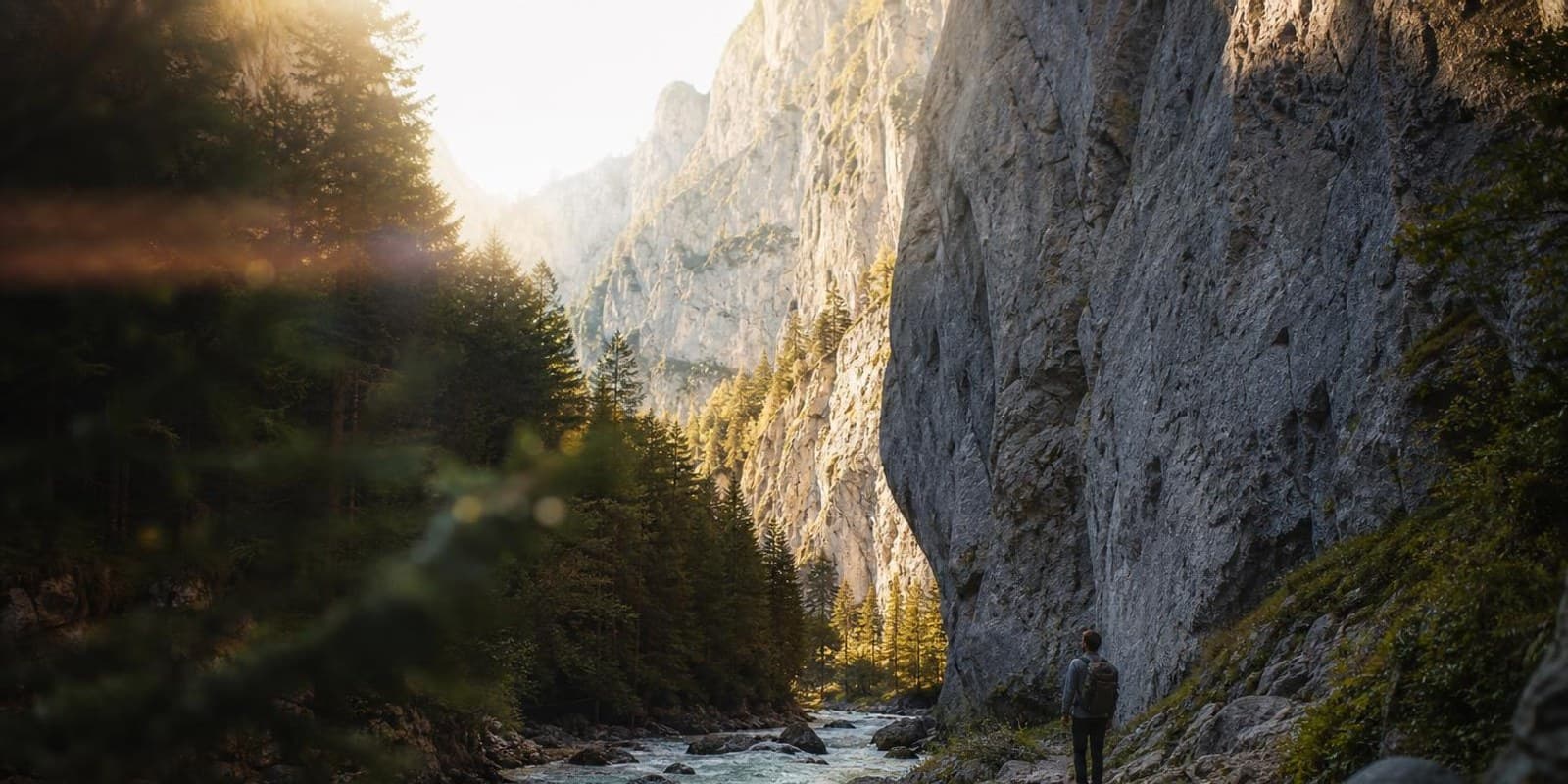 Wanderer geht am Ufer eines wilden Flusses zwischen hohen hellen Kalkwaenden entlang.