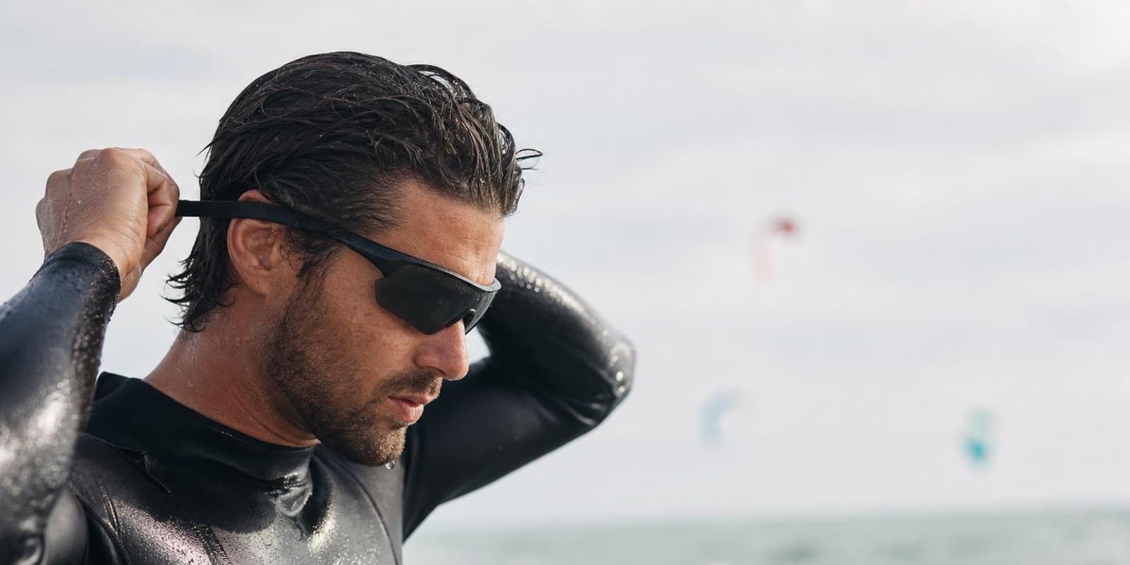 Kitesurfer befestigt eine Sportbrille mit Band, im Hintergrund Kites ueber dem Meer.