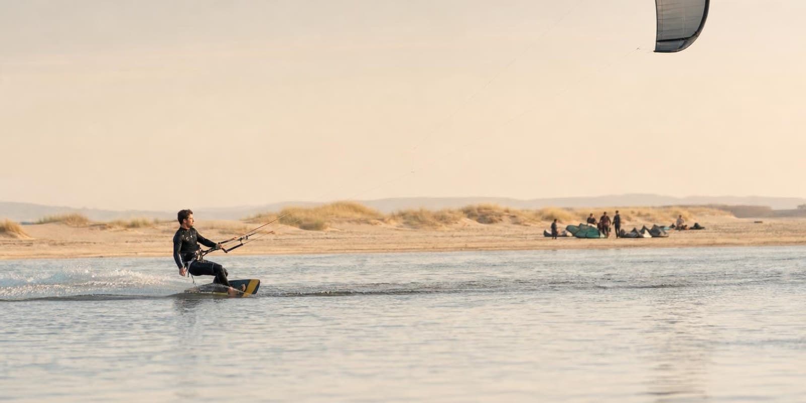 Kiter gleitet mit grossem Schirm ueber spiegelglattes Wasser, waehrend andere am Strand auf Wind warten.