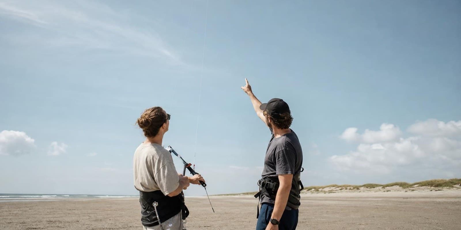 Schueler blickt am Strand zu einem hoch stehenden Uebungskite hinauf, der Himmel dominiert das Bild.
