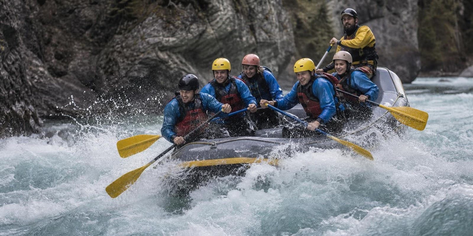 Raft-Crew mit Helmen und Schwimmwesten paddelt durch eine schaeumende Wildwasserwelle.