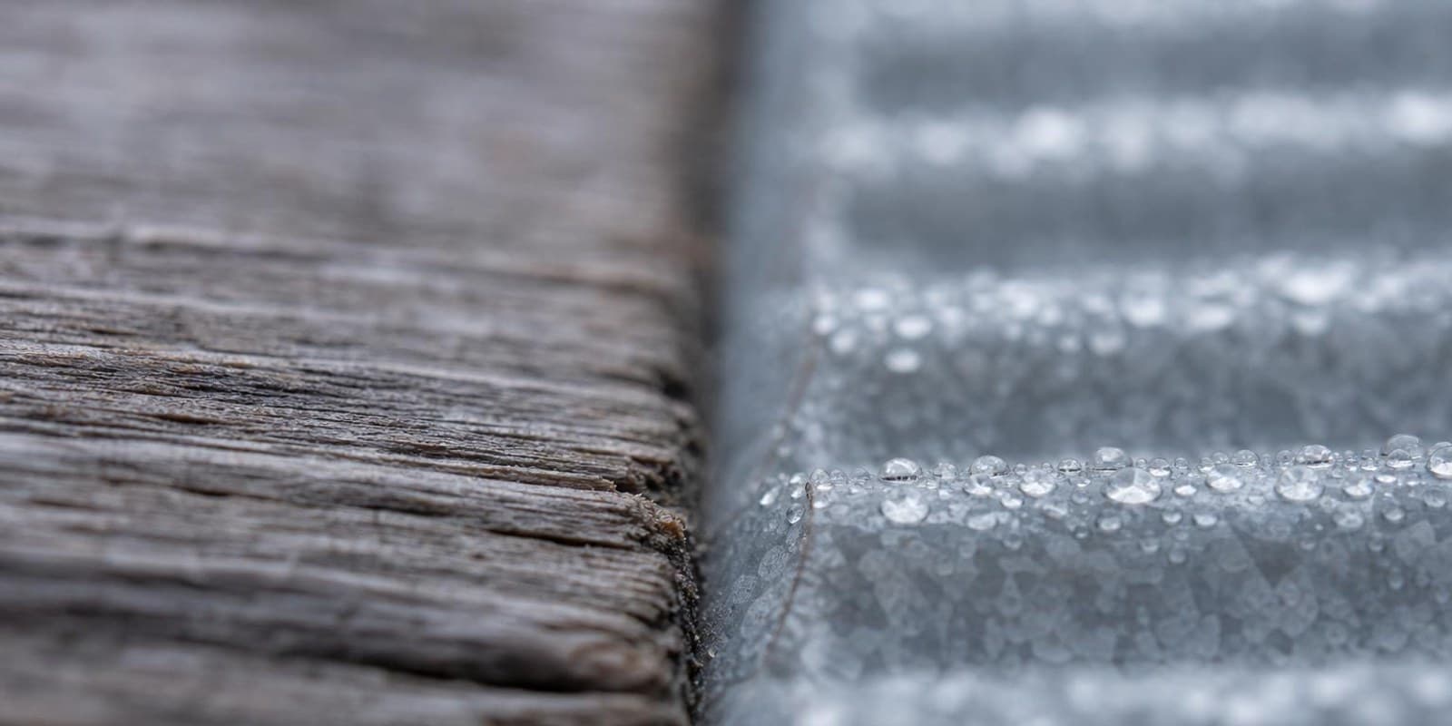 Nahaufnahme einer verwitterten Holzbohle neben einer verzinkten Metallwand mit Tautropfen.