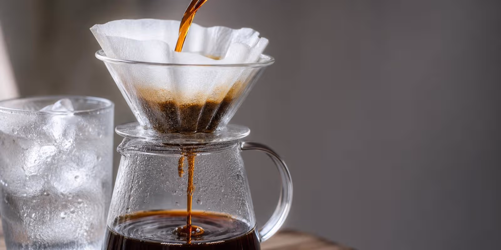 Cold Brew laeuft durch einen Papierfilter in eine Glaskaraffe, daneben ein Glas mit Eis.