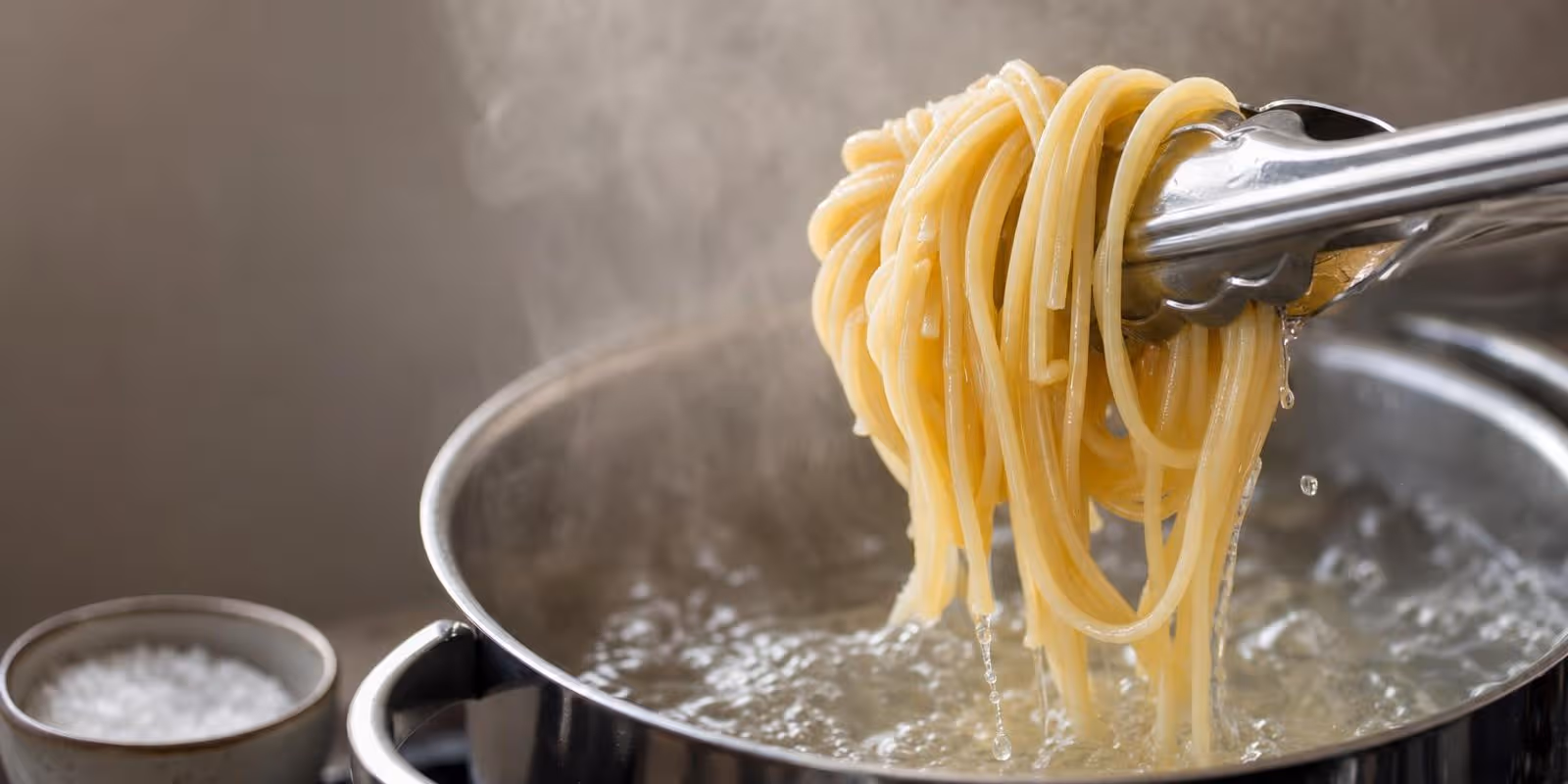 Eine Zange hebt glaenzende Spaghetti aus sprudelnd kochendem Salzwasser, Dampf steigt auf.