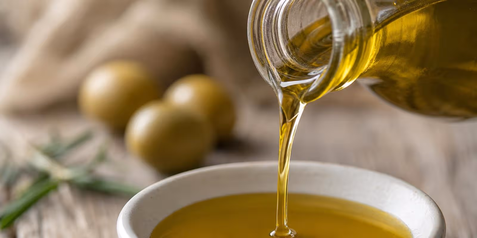 Olivenoel fliesst in eine kleine weisse Schale, daneben Oliven und Rosmarin.