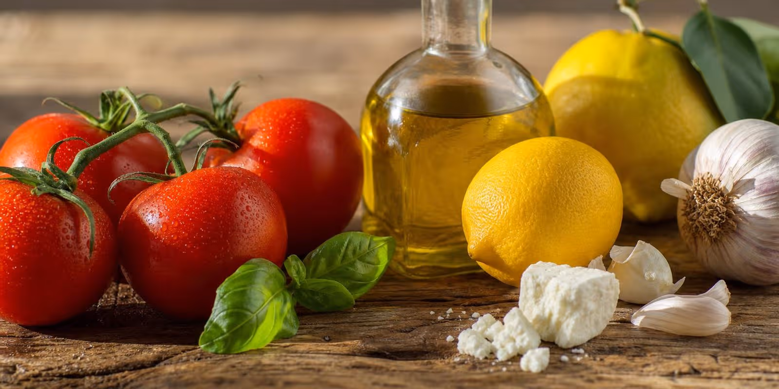 Tomaten, Olivenoel, Zitronen und Kraeuter liegen auf einem sonnenbeschienenen Holztisch.