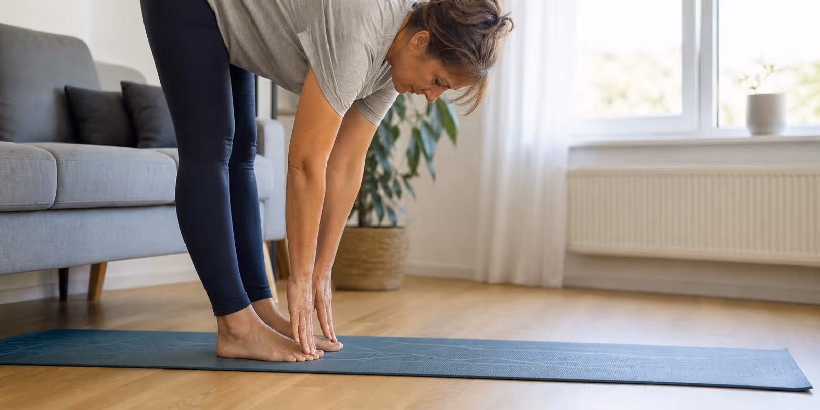 Frau testet auf einer Matte im Wohnzimmer mit dem Zehenspitzen-Test ihre Beweglichkeit.