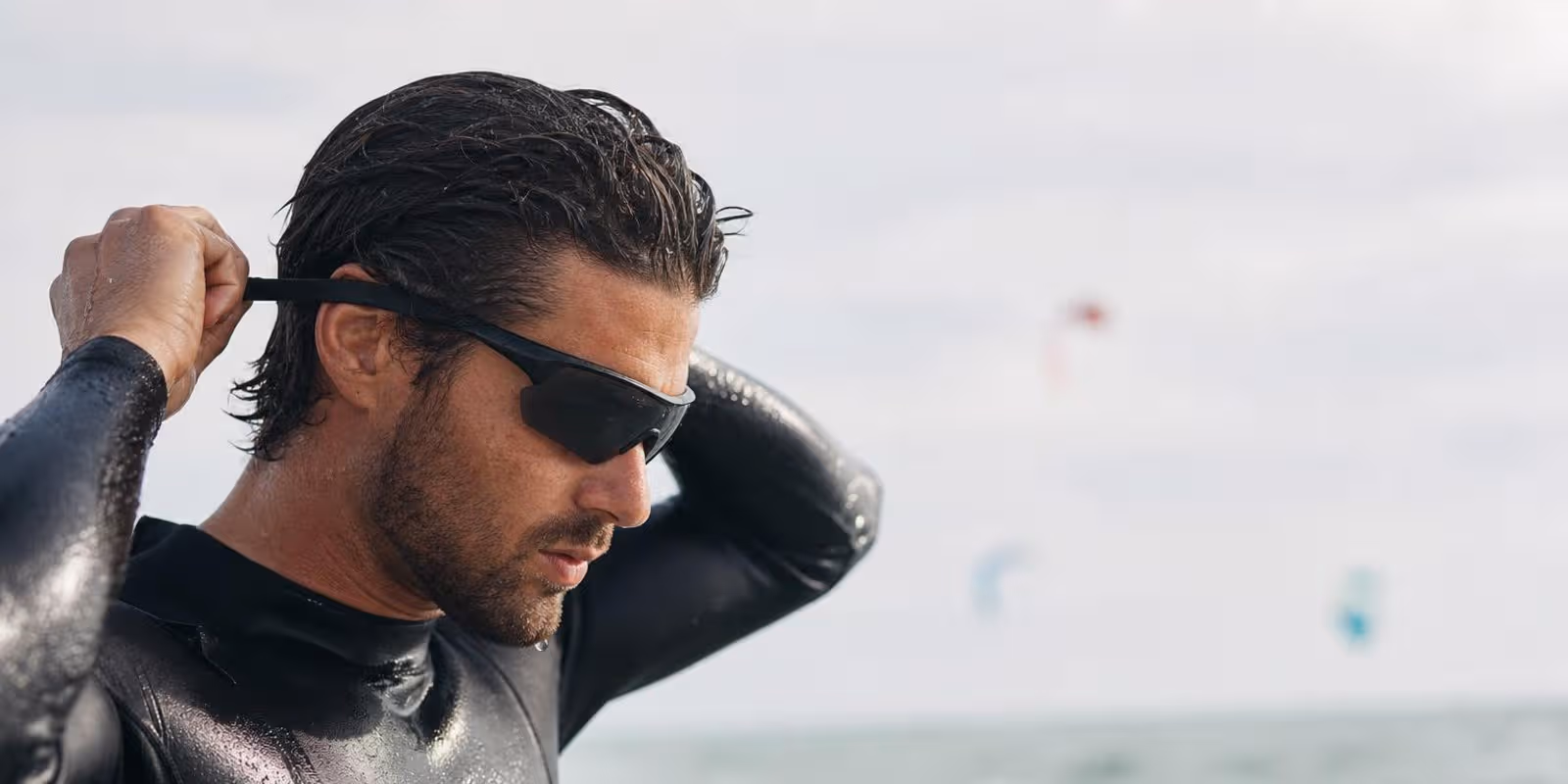 Kitesurfer befestigt eine Sportbrille mit Band, im Hintergrund Kites ueber dem Meer.