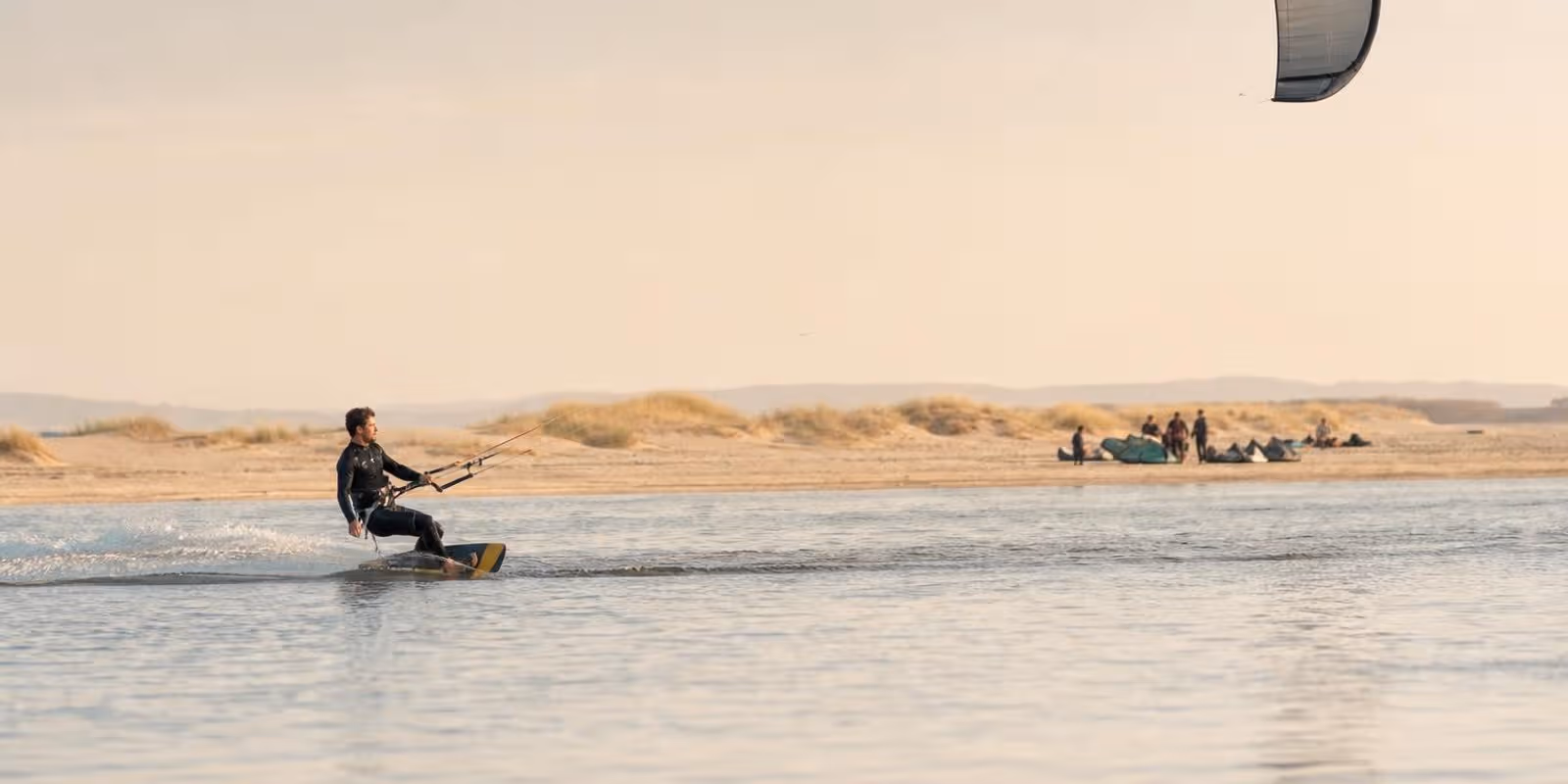 Kiter gleitet mit grossem Schirm ueber spiegelglattes Wasser, waehrend andere am Strand auf Wind warten.