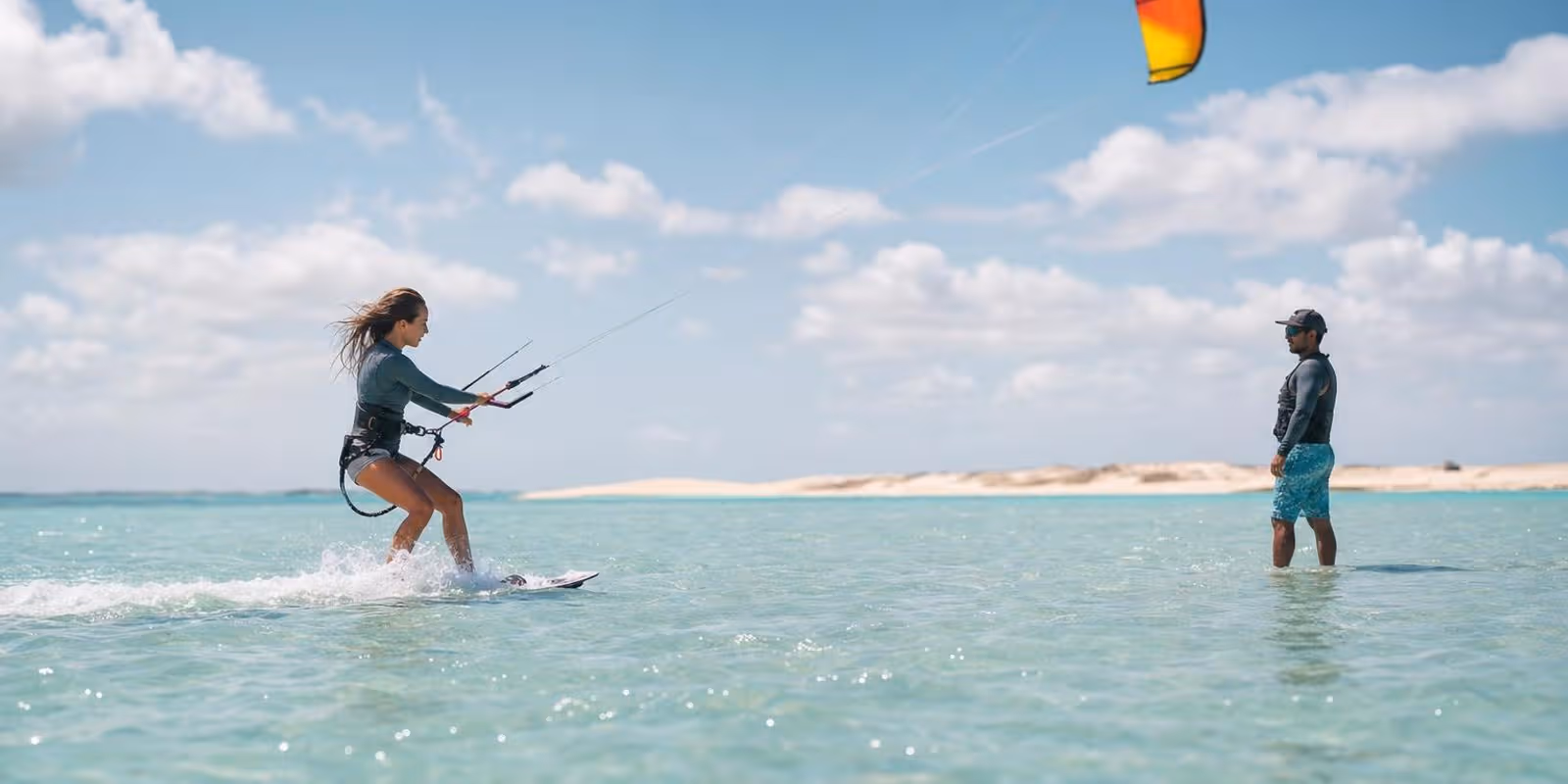 Ein Kite-Anfaenger faehrt durch flaches tuerkises Wasser, waehrend ein Lehrer in der Naehe zusieht.