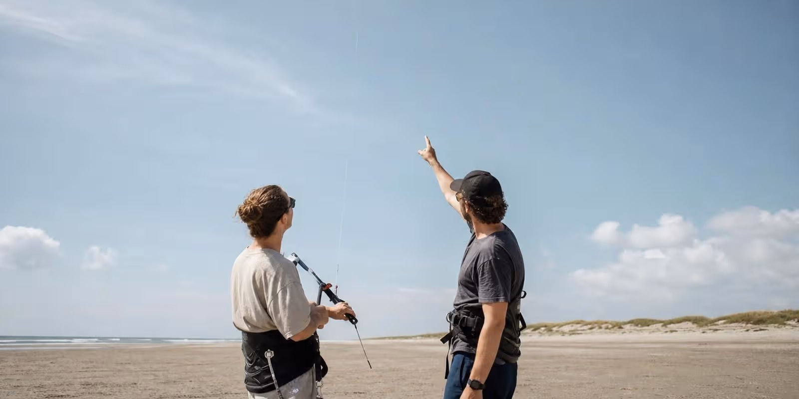 Schueler blickt am Strand zu einem hoch stehenden Uebungskite hinauf, der Himmel dominiert das Bild.