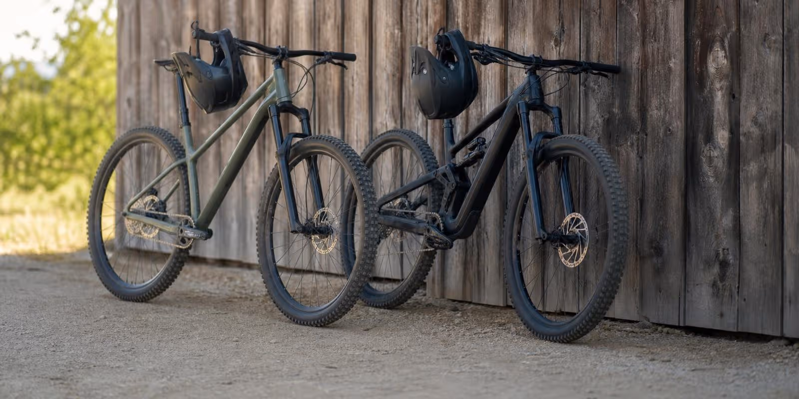 Hardtail und Fully lehnen zum Vergleich nebeneinander an einer Holzwand, Helme am Lenker.