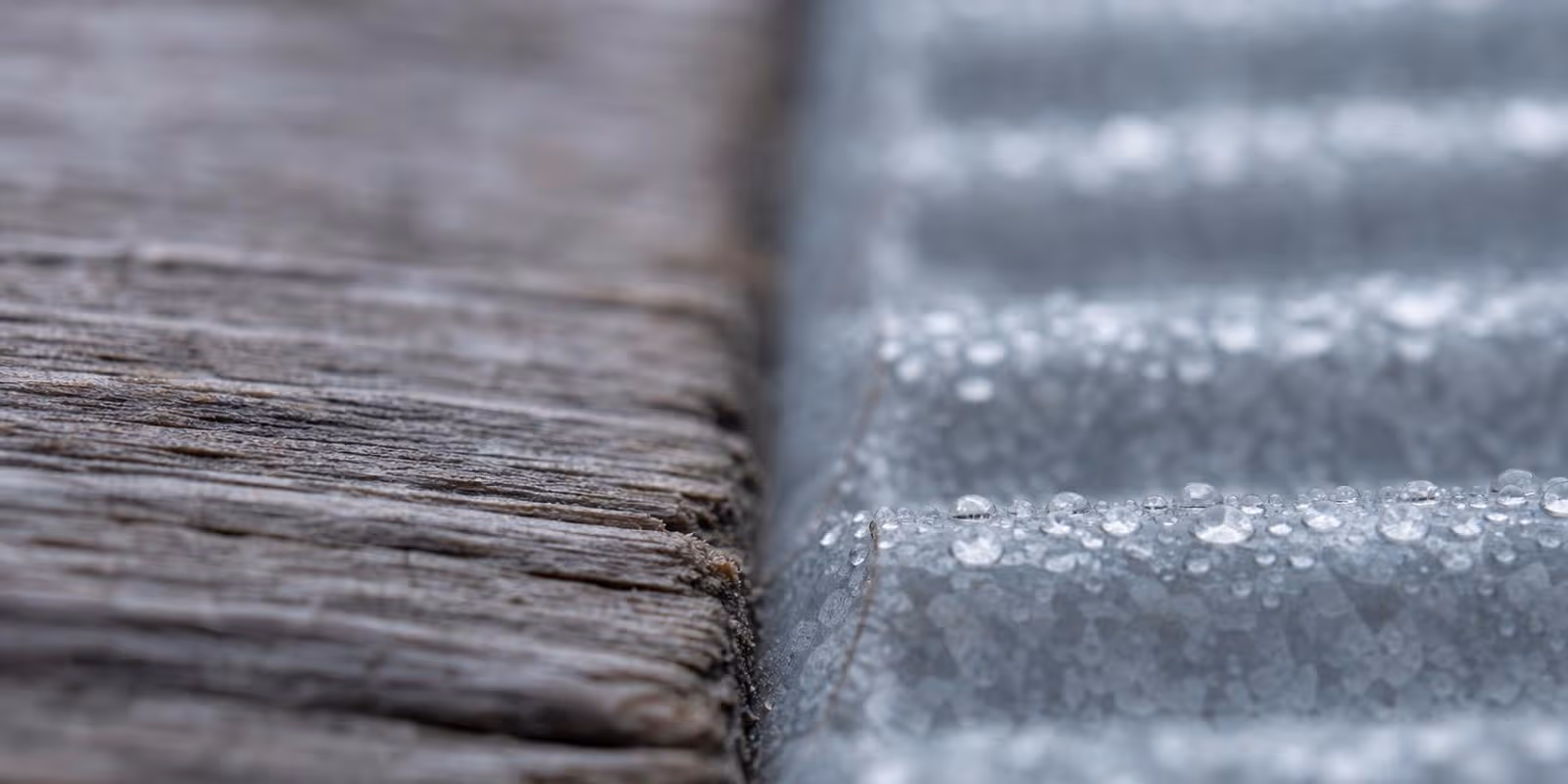 Nahaufnahme einer verwitterten Holzbohle neben einer verzinkten Metallwand mit Tautropfen.