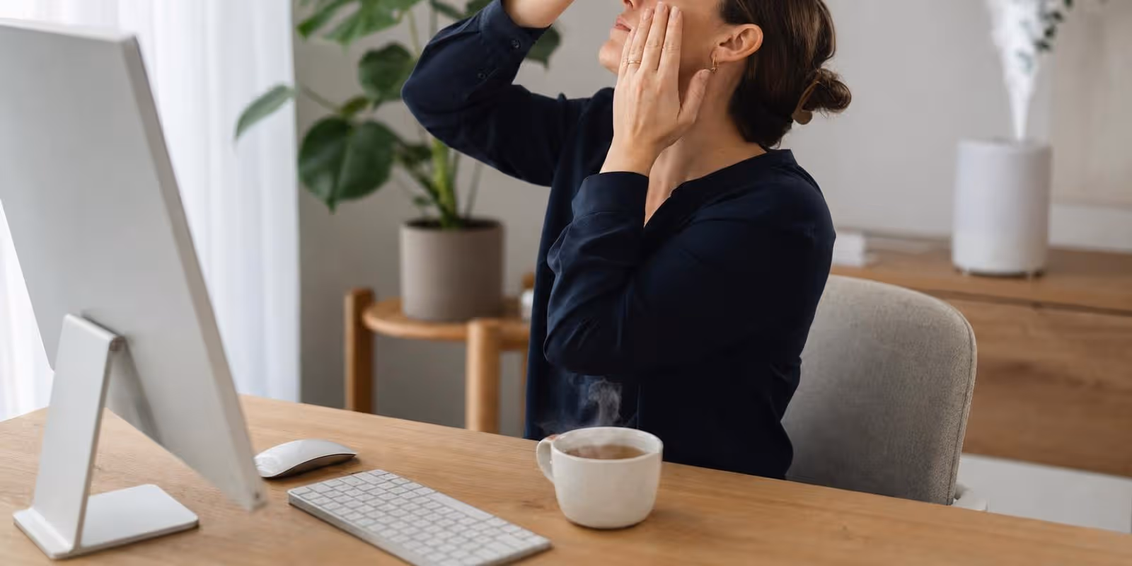 Eine Frau traeufelt sich am Schreibtisch entspannt einen Augentropfen ins Auge, daneben dampft eine Teetasse.