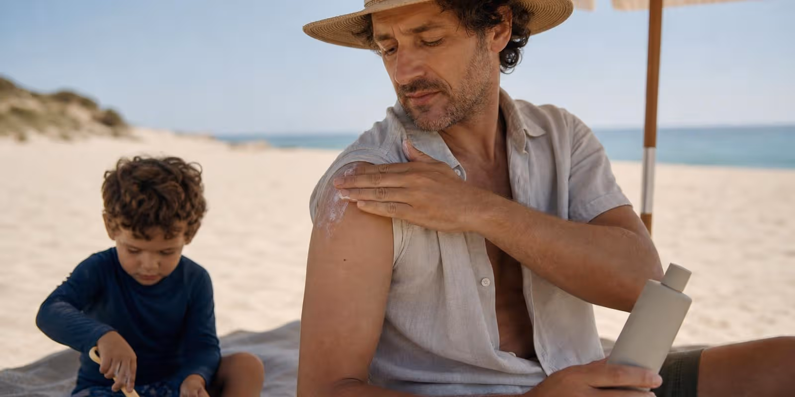 Mann cremt sich im Schatten eines Sonnenschirms am Strand mit Sonnencreme ein.