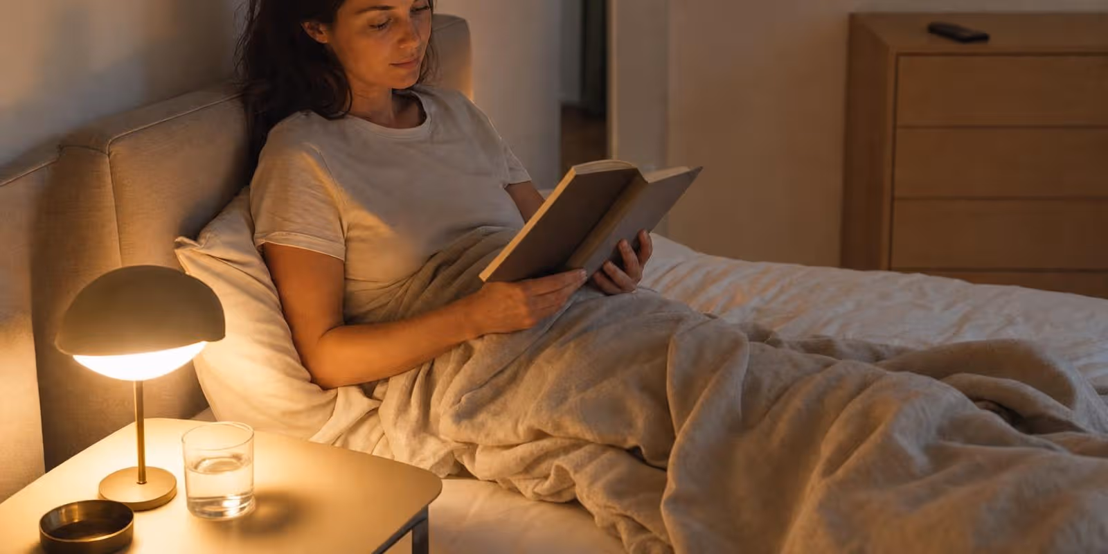 Eine Frau liest abends im Bett ein Buch im warmen Licht der Nachttischlampe, das Handy liegt entfernt auf der Kommode.
