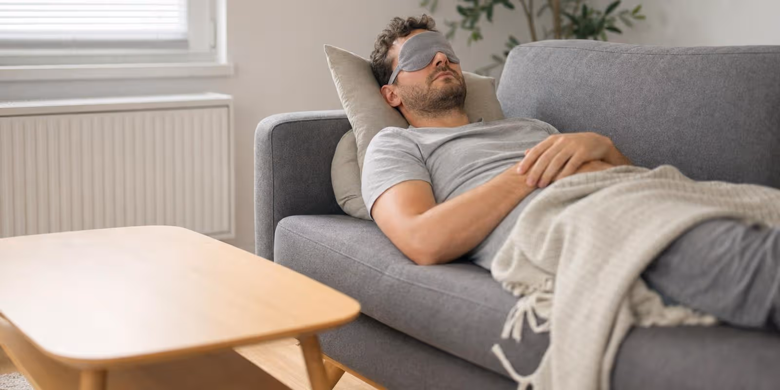 Ein Mann haelt mit Schlafmaske und leichter Decke einen kurzen Mittagsschlaf auf dem Sofa.