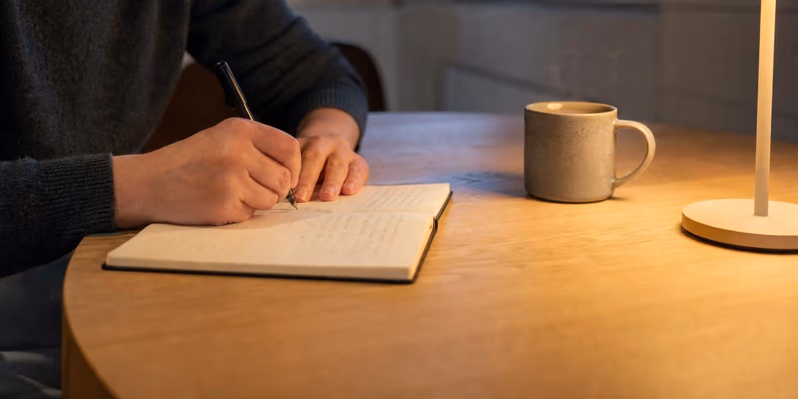 Haende schreiben abends bei Lampenlicht in ein einfaches Notizbuch, daneben steht eine Tasse Tee.