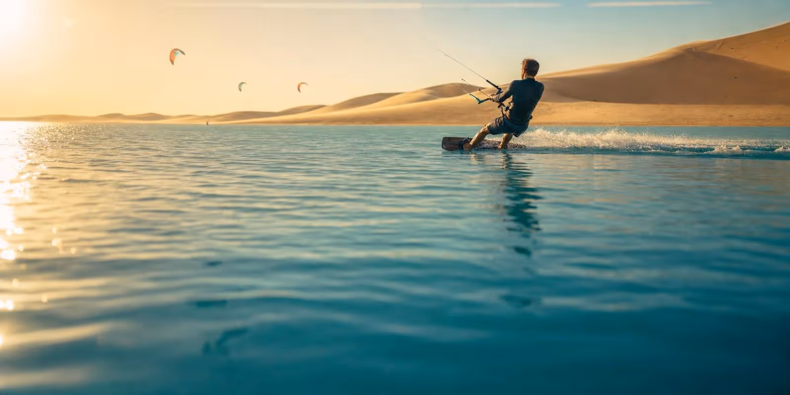 Kitesurfer gleiten ueber eine flache tuerkise Lagune, dahinter leuchten die Sandduenen der Sahara.