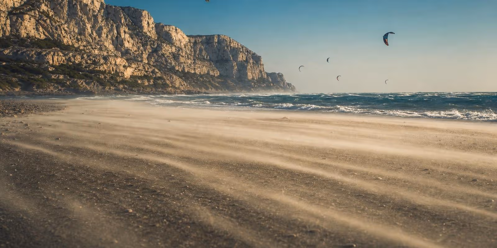 Sturmgepeitschter Strand bei Leucate, Sand weht flach ueber den Boden und viele Kites rasen ueber Schaumkronen.