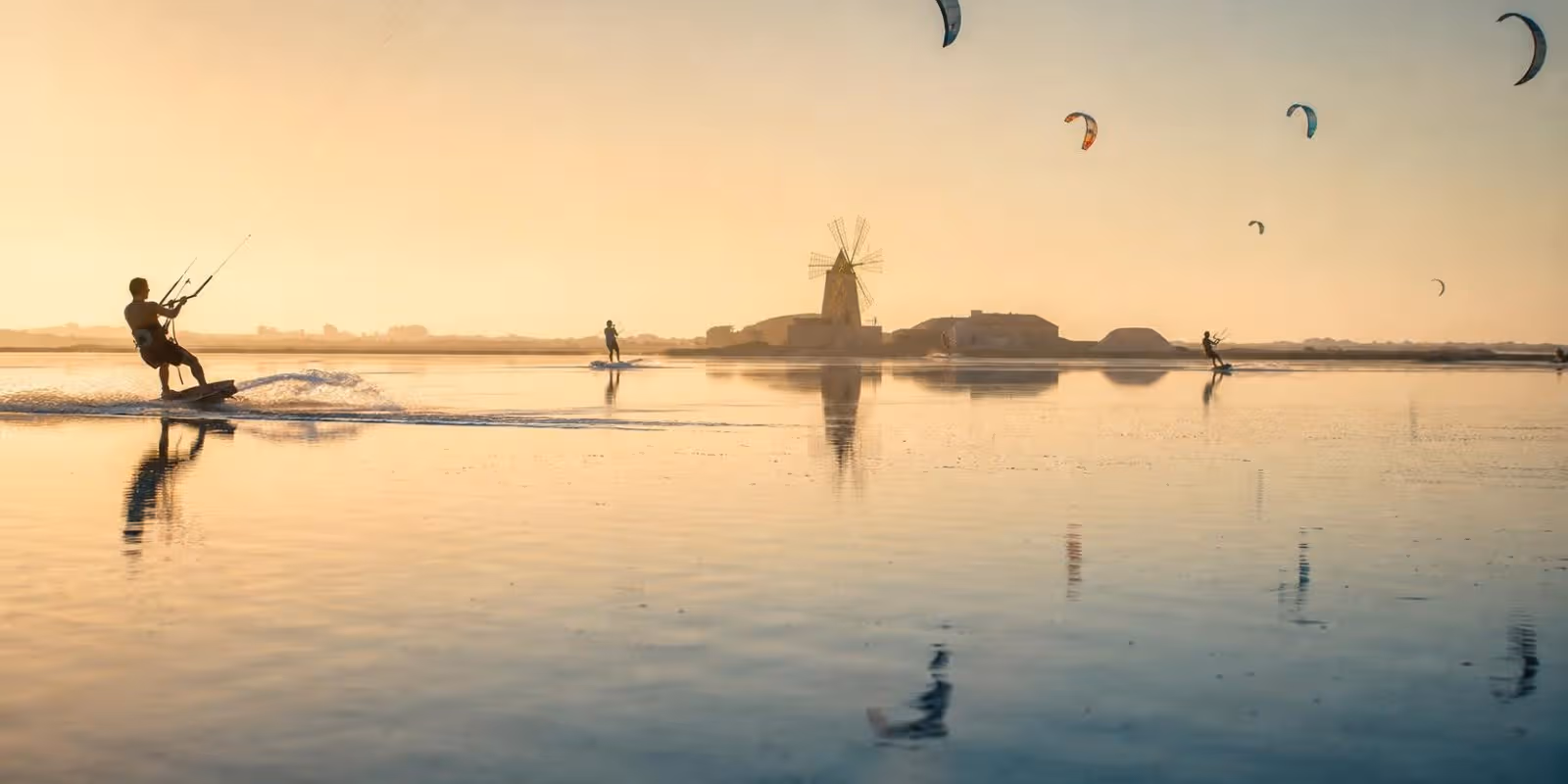 Kniehohe Lagune auf Sizilien mit Kitesurfern, am Ufer die Silhouette einer alten Windmuehle.