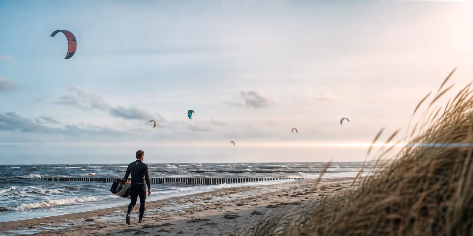 Kitesurfer traegt sein Board am Ostseestrand mit Buhnen entlang, Kites am Himmel.