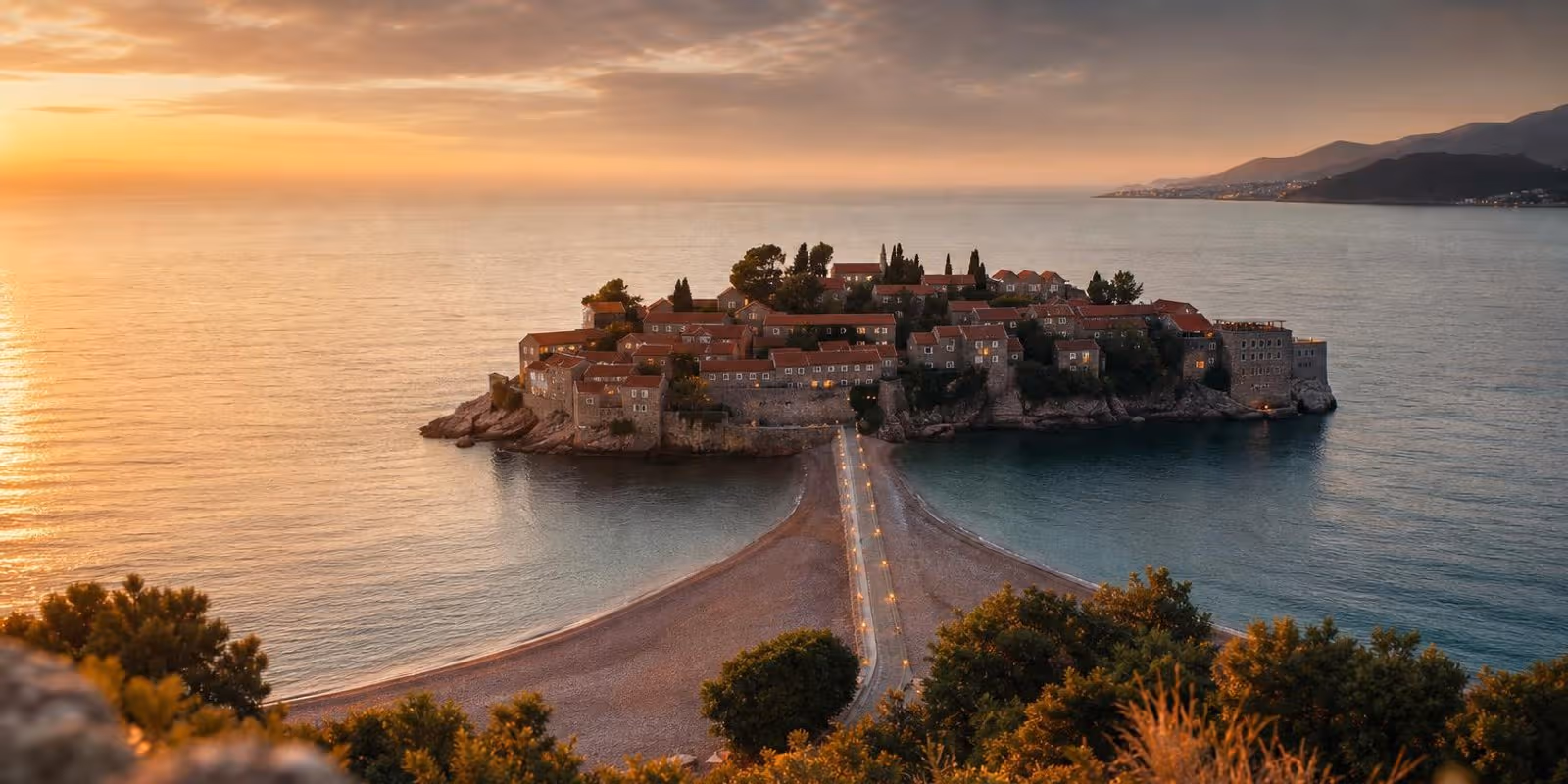 Inselort Sveti Stefan mit Damm und rosafarbenen Kiesstraenden im Abendlicht.