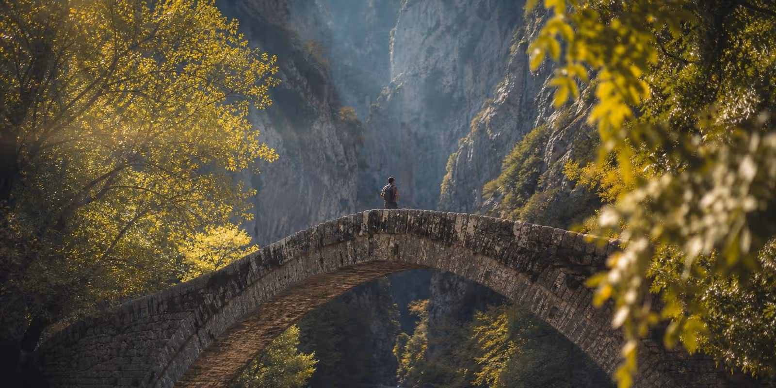 Wanderer ueberquert eine alte Steinbogenbruecke in Zagori, dahinter oeffnet sich die Schlucht.