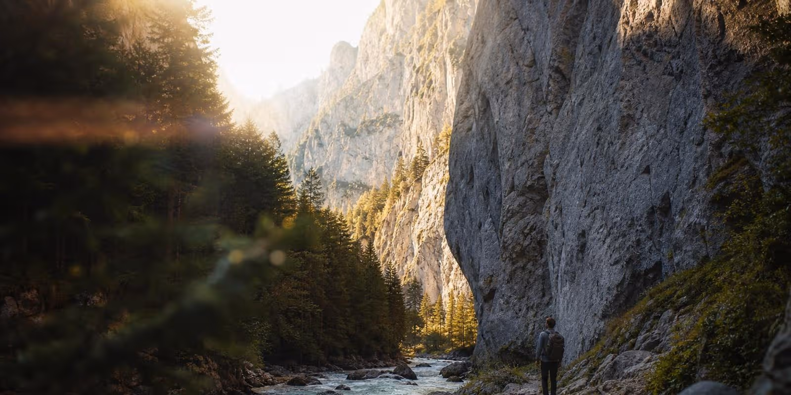 Wanderer geht am Ufer eines wilden Flusses zwischen hohen hellen Kalkwaenden entlang.