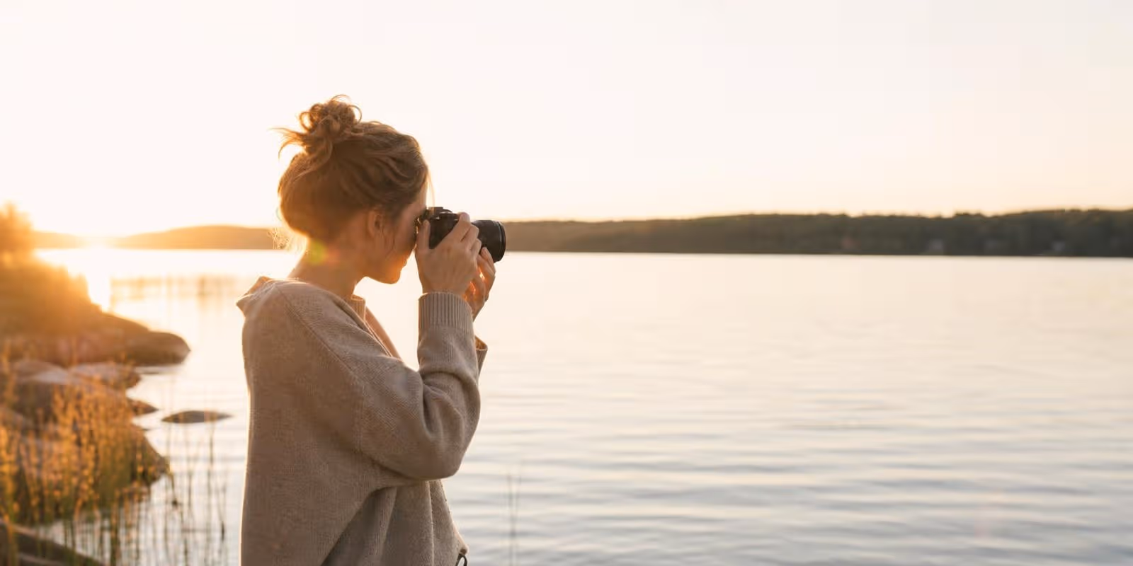 Fotografin haelt am Seeufer im Abendlicht die Kamera ans Auge und dreht am Objektivring.