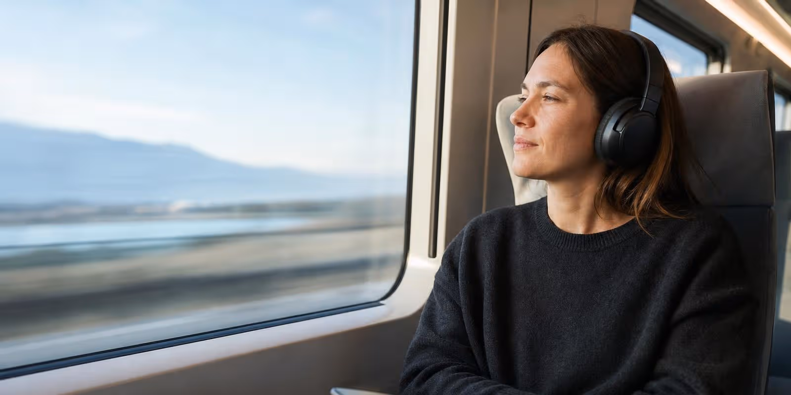 Pendler sitzt mit Over-Ear-Kopfhoerern am Zugfenster, draussen zieht die Landschaft vorbei.