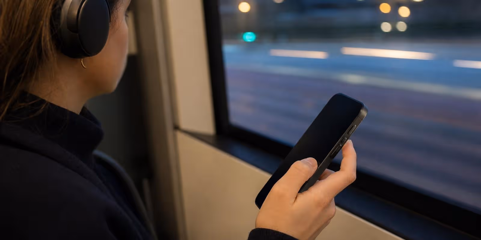 Junge Frau mit Kopfhoerern in der Bahn haelt den Daumen ueber dem Lautstaerkeregler ihres Handys.