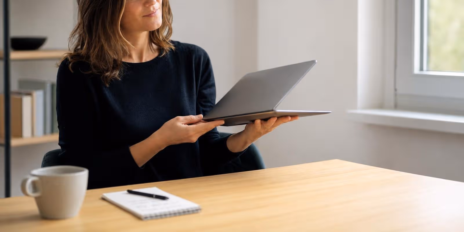 Eine Frau wiegt nachdenklich einen flachen Laptop in den Haenden, daneben liegt ein Notizblock.