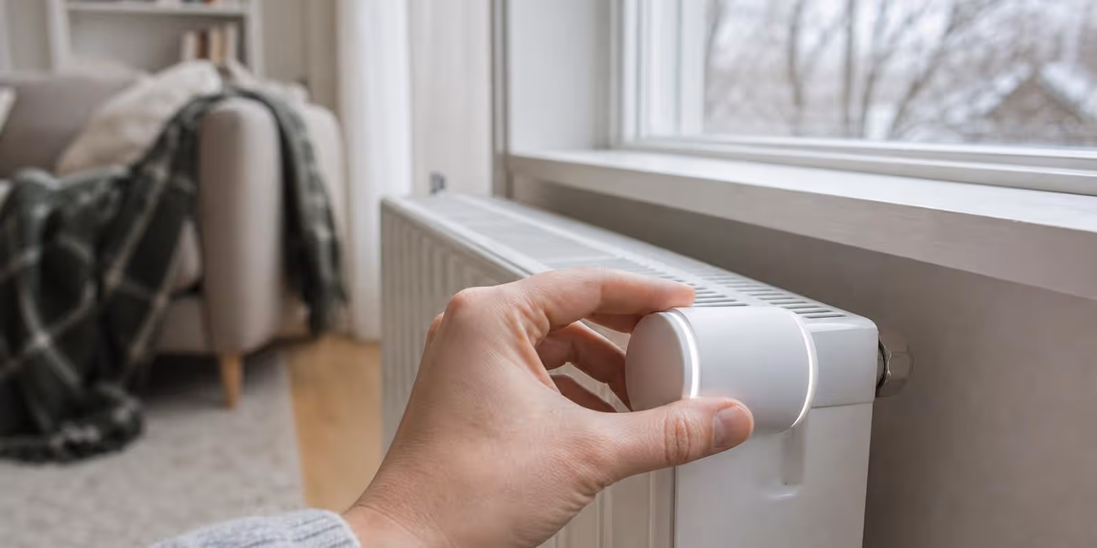 Hand stellt ein smartes Thermostat am Heizkoerper unter dem Fenster ein.