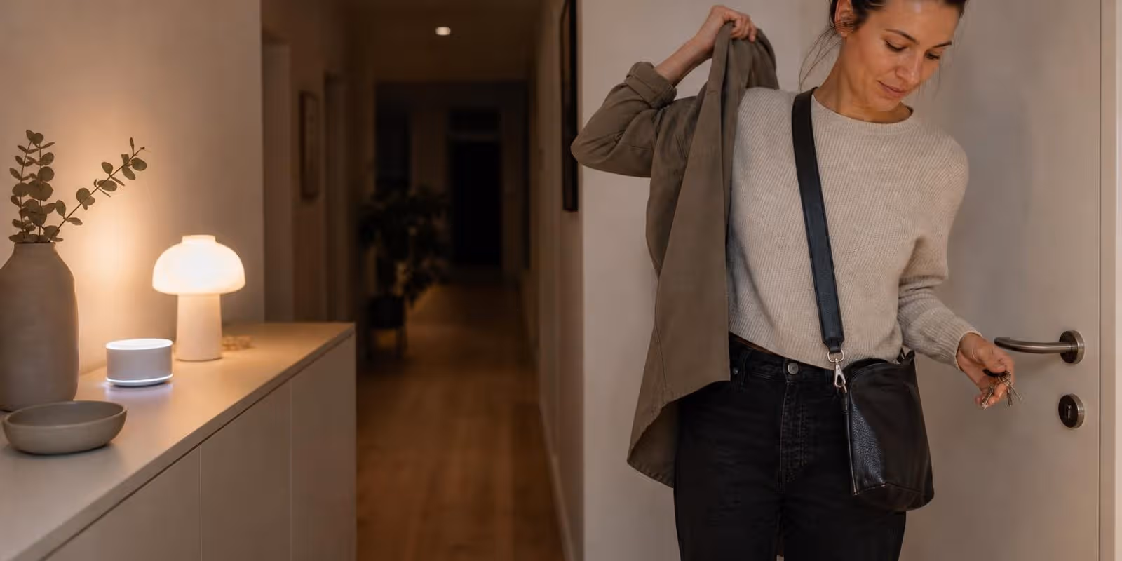 Person zieht abends im Flur die Jacke an, waehrend Lampe und Lautsprecher auf der Kommode leuchten.